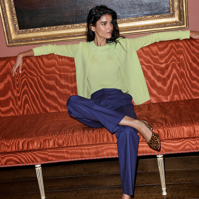A woman sits on a rust-orange velvet sofa in a styled interior, wearing a light lime green draped long-sleeve top and navy blue trousers, with leopard-print shoes.