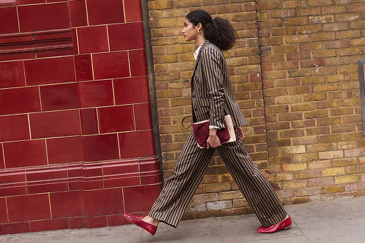 A woman walks in profile wearing a brown and beige pinstripe blazer and matching wide-leg trousers, holding a burgundy clutch, with red shoes, set against a split red tile and brick wall background.