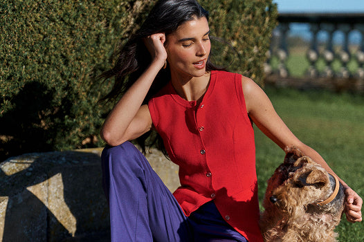A woman wearing a sleeveless red top with buttons and purple pants sits outdoors next to a dog, with greenery and a stone wall in the background.