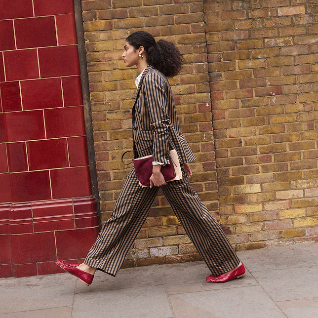A woman walks in profile wearing a brown and beige vertically striped blazer and matching wide-leg trousers, holding a burgundy clutch, with red shoes, set against a brick wall and red tiled wall background.