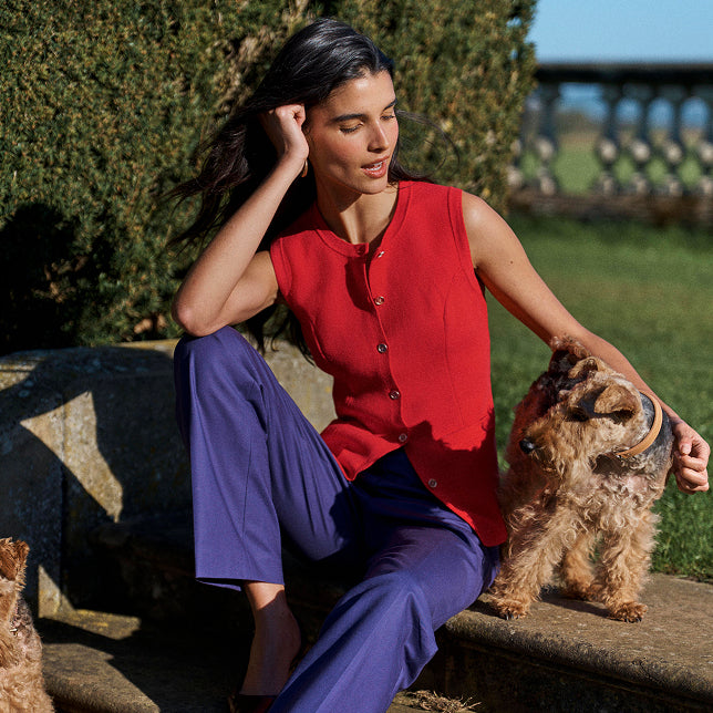 A woman sits outdoors on a stone bench wearing a sleeveless red top and purple pants, with a small brown dog beside her against a garden background.