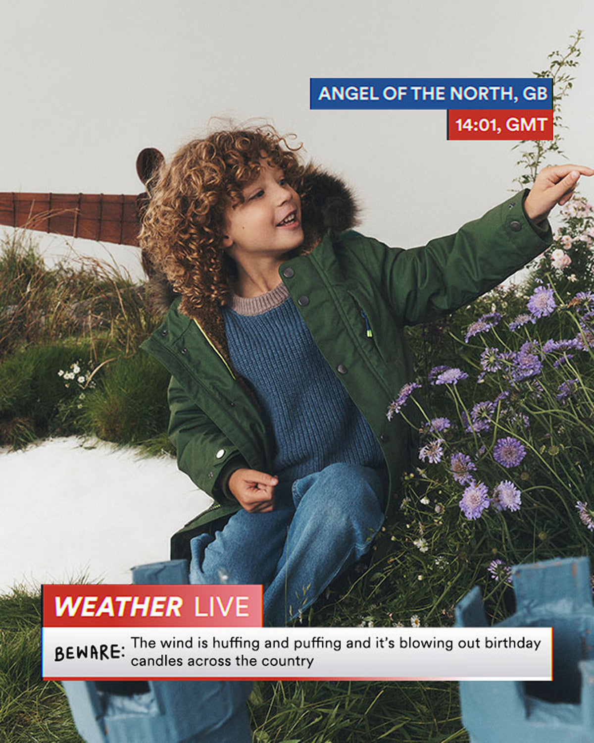 A child wearing a green parka with a fur-trimmed hood over a blue knit sweater and jeans, sitting outdoors in grass with purple flowers. Text overlays read 'ANGEL OF THE NORTH, GB', '14:01, GMT', 'WEATHER LIVE', and 'Beware: The wind is huffing and puffing and it's blowing out birthday candles across the country.