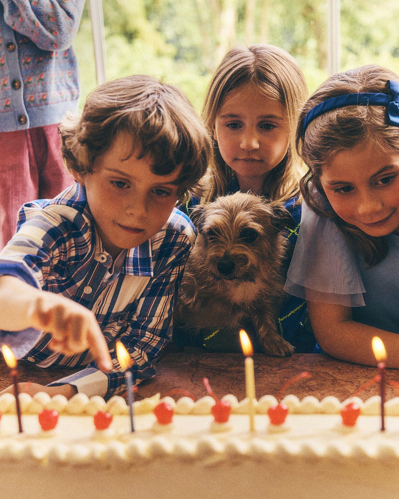 Three children and a small dog gathered around a birthday cake with lit candles in a warm indoor setting.