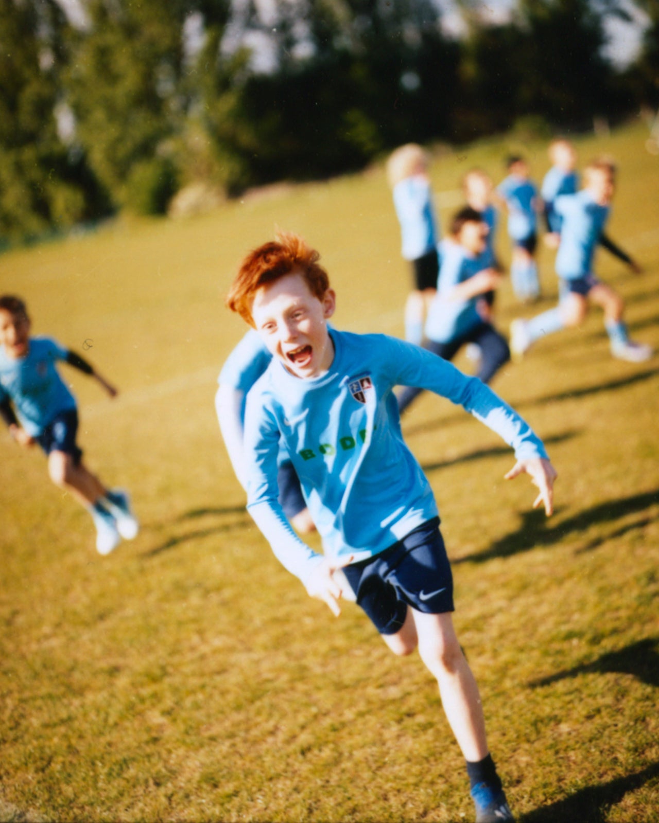 Children playing soccer on a grassy field with trees in the background