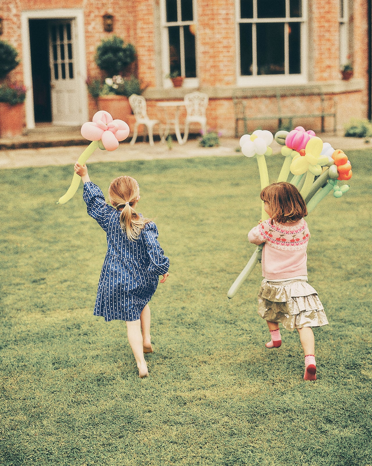 Two young girls running across a green lawn in front of a brick house, each holding a bouquet of oversized balloon flowers in pastel colors.