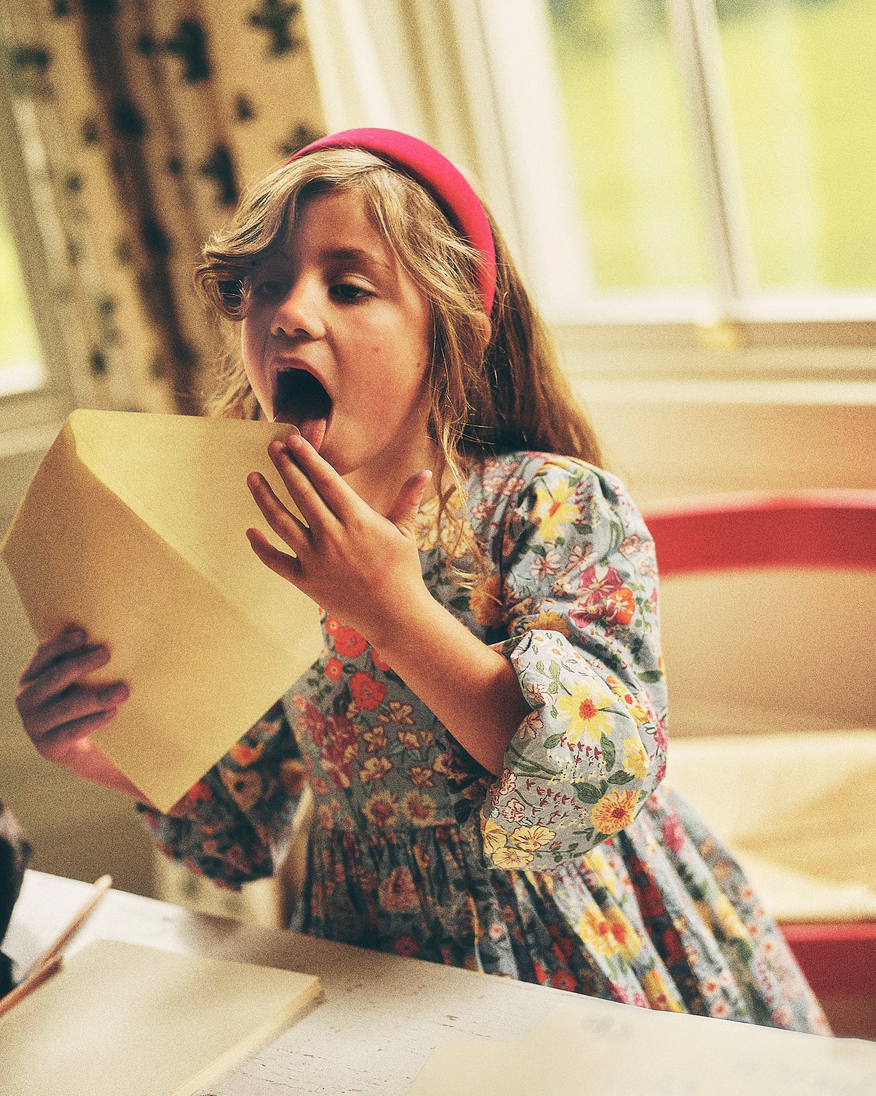 A young girl with a pink headband and a colorful floral dress holds a large yellow envelope, looking surprised with her mouth open in a brightly lit room.