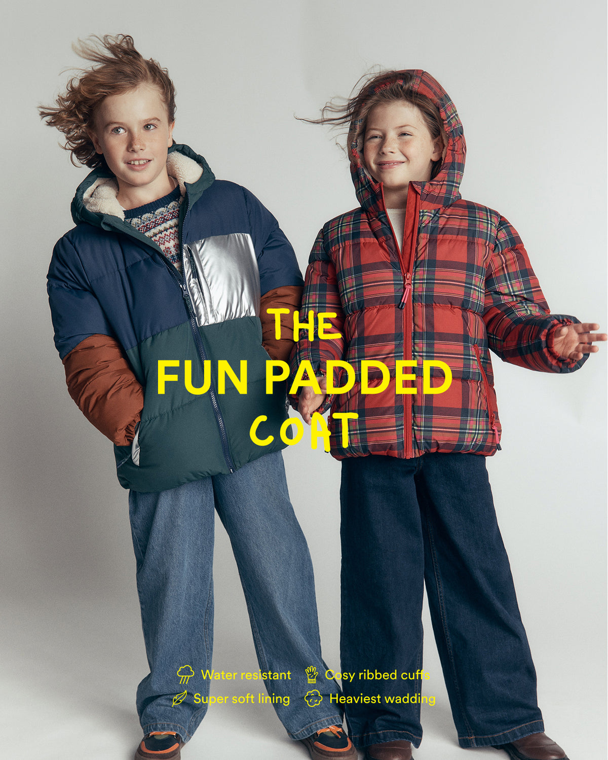 Two children modeling padded winter coats against a light gray studio background. The child on the left wears a navy, green, and brown color-blocked coat with a silver panel, and the child on the right wears a red and black plaid hooded coat.