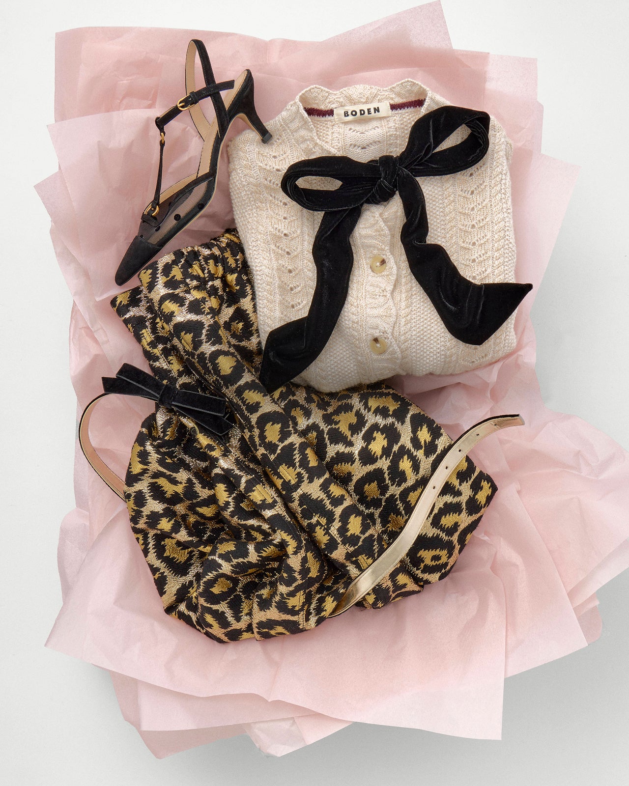 Leopard print handbag, black high-heeled shoes, and a white cardigan with a bow on a pink surface.