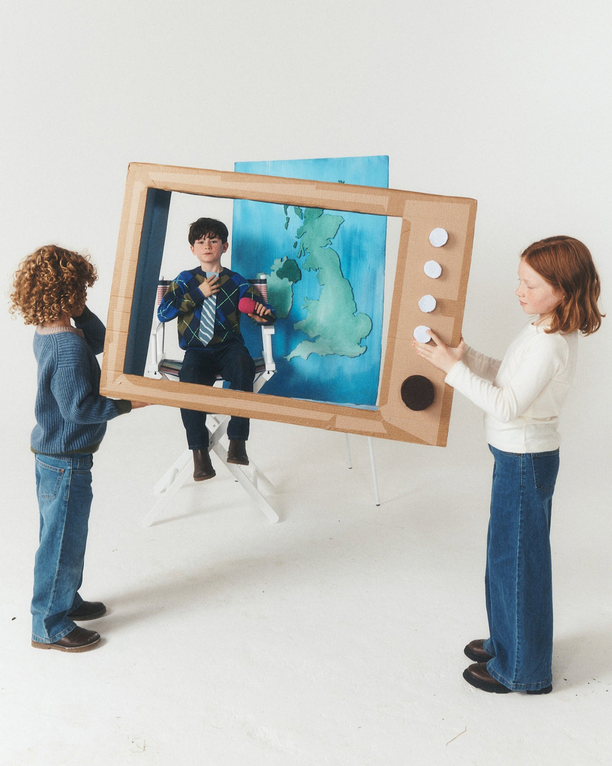 Three children are playing with a large cardboard toy television set in a bright white studio. The TV screen shows a painted map of the United Kingdom, and one child is seated inside the TV as if watching a program.