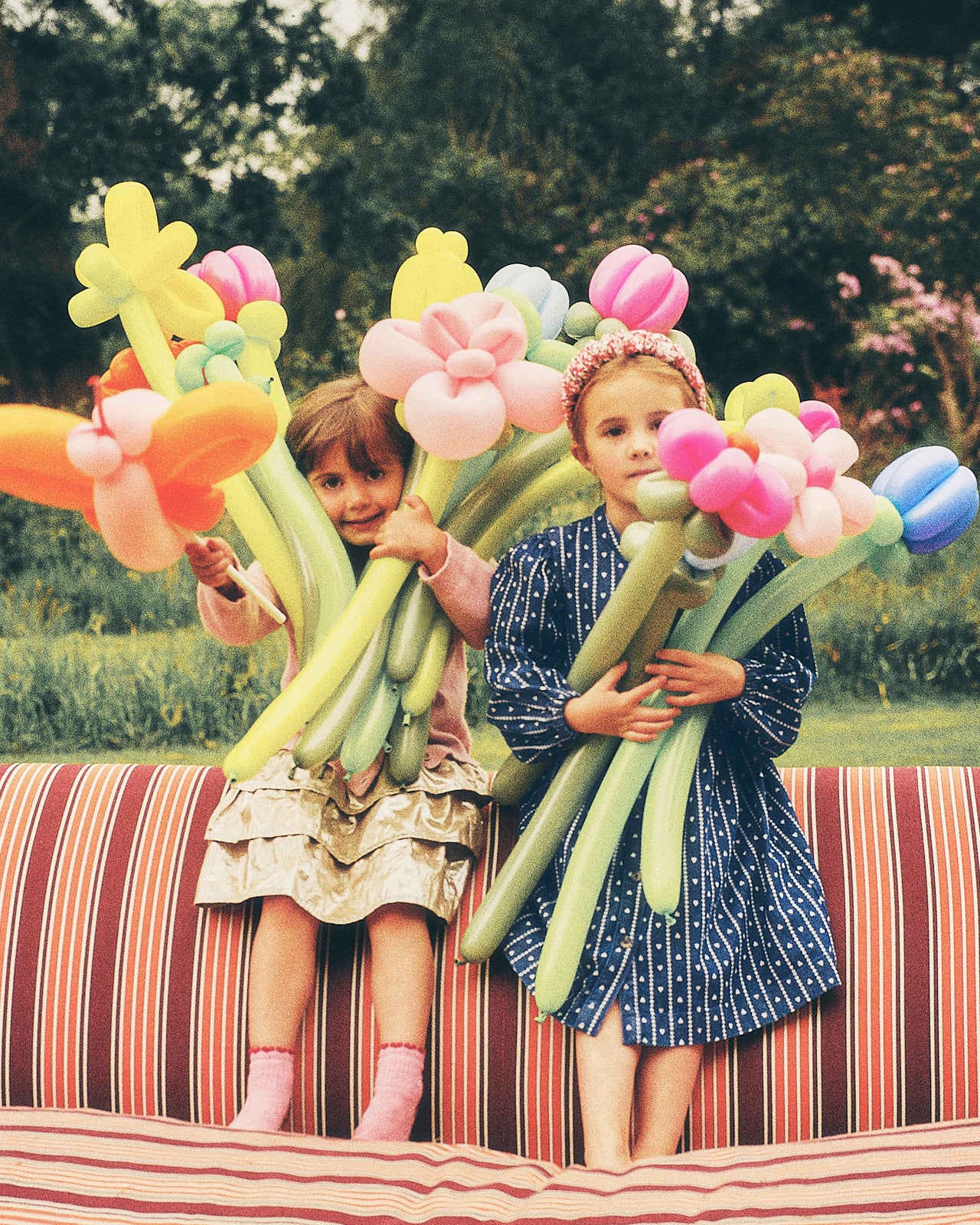 Two young girls sitting on a striped outdoor bench holding large balloon flower sculptures in a park setting.