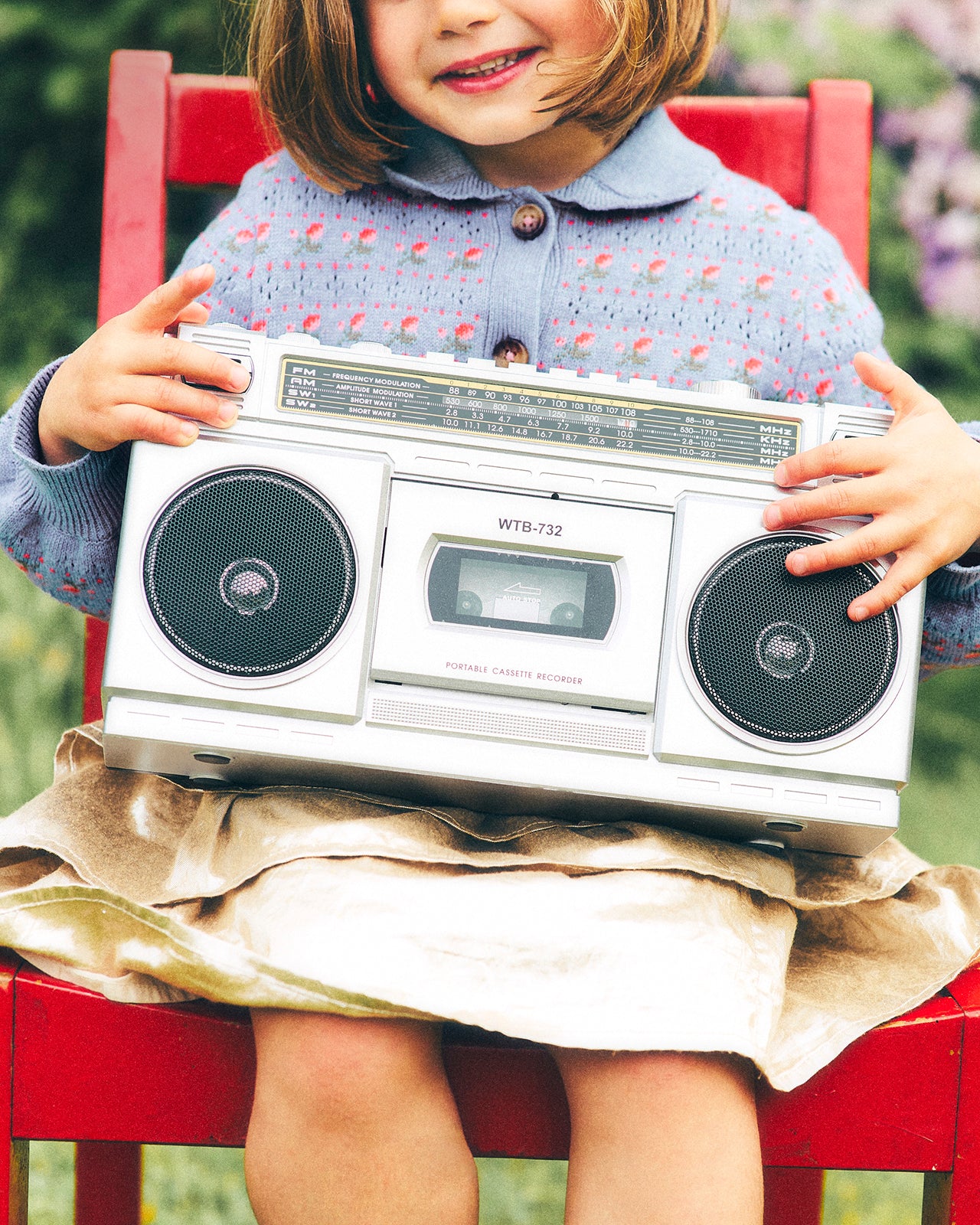 A young girl in a blue patterned sweater and beige skirt sits on a red wooden chair, holding a vintage silver cassette boombox radio with black speakers.