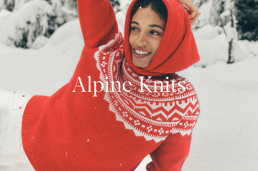Person wearing a red knitted outfit with white patterns, lying on a snowboard in a snowy landscape. 'Alpine Knits' text overlay.