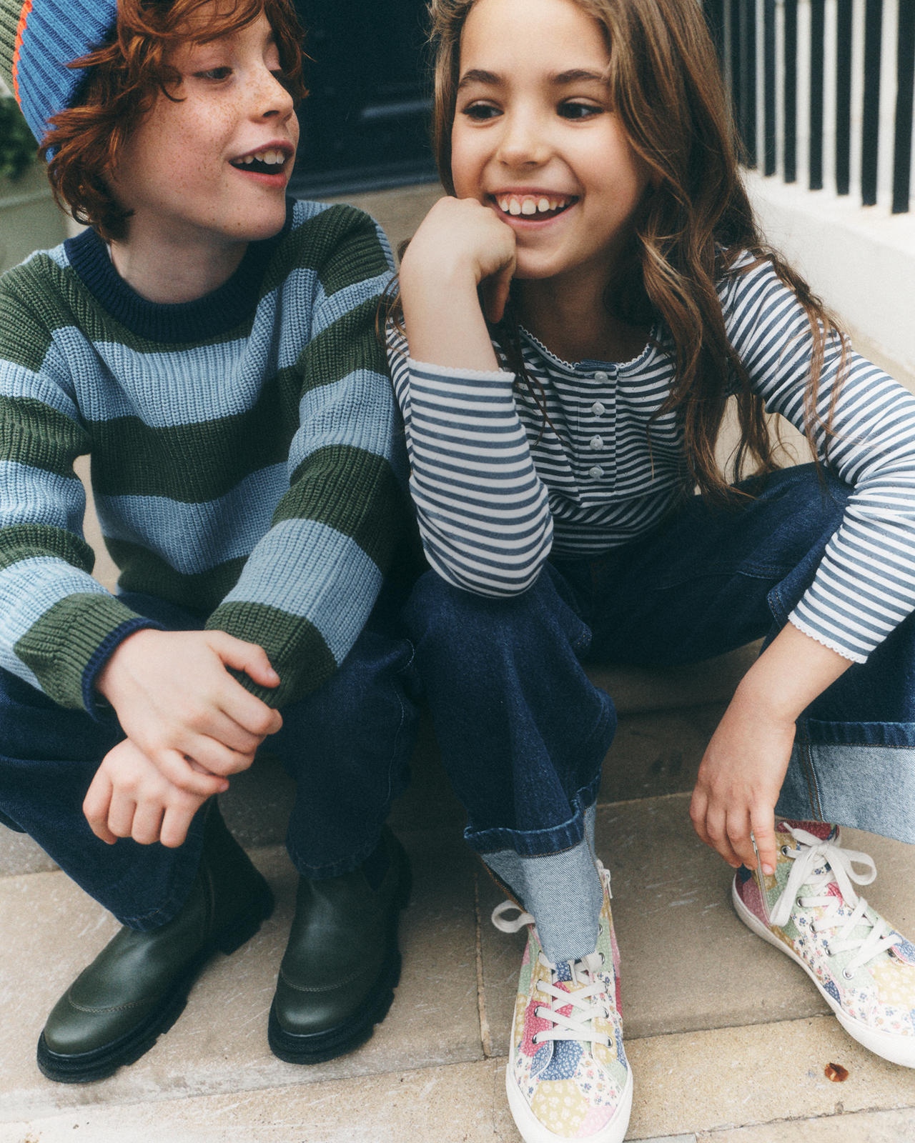 Two children sitting together on outdoor steps, wearing striped knit sweaters and casual jeans. The boy wears a blue and green striped sweater with dark boots, and the girl wears a blue and white striped long-sleeve top with multicolored sneakers.