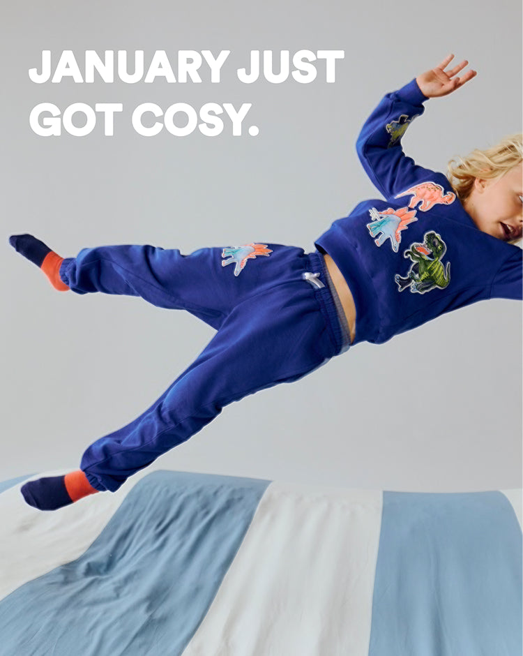 Child wearing a blue outfit with dinosaur designs, jumping on a bed with a light gray background.