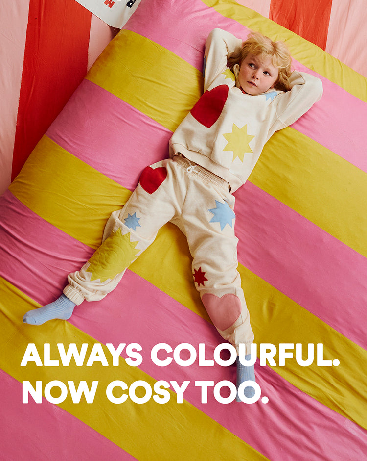 Child wearing a colorful outfit lying on a striped pink and yellow blanket with text 'Always colourful. Now cosy too.'