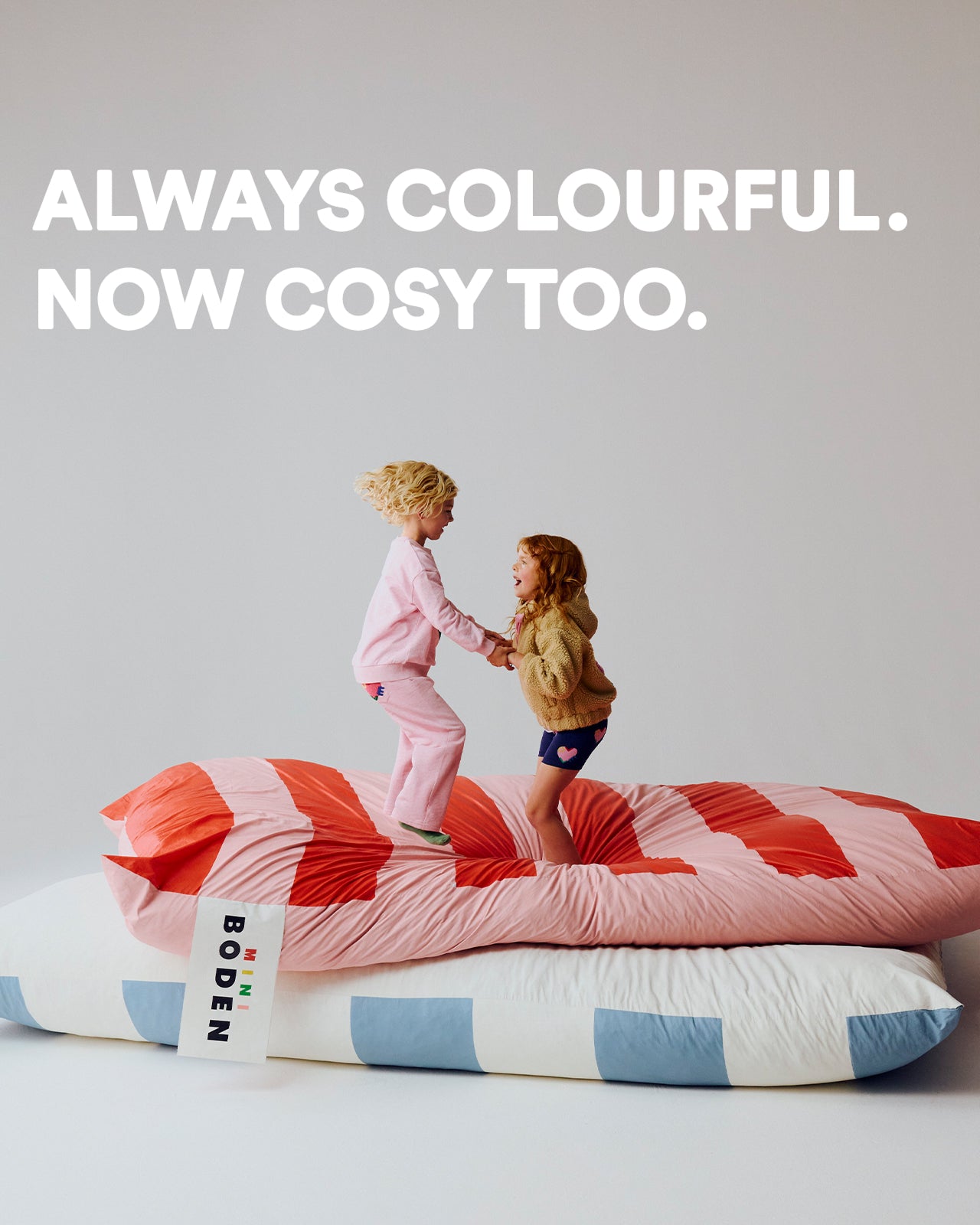 Two children on a bed with a colorful quilt, text 'Always Colourful. Now Cosy Too.'