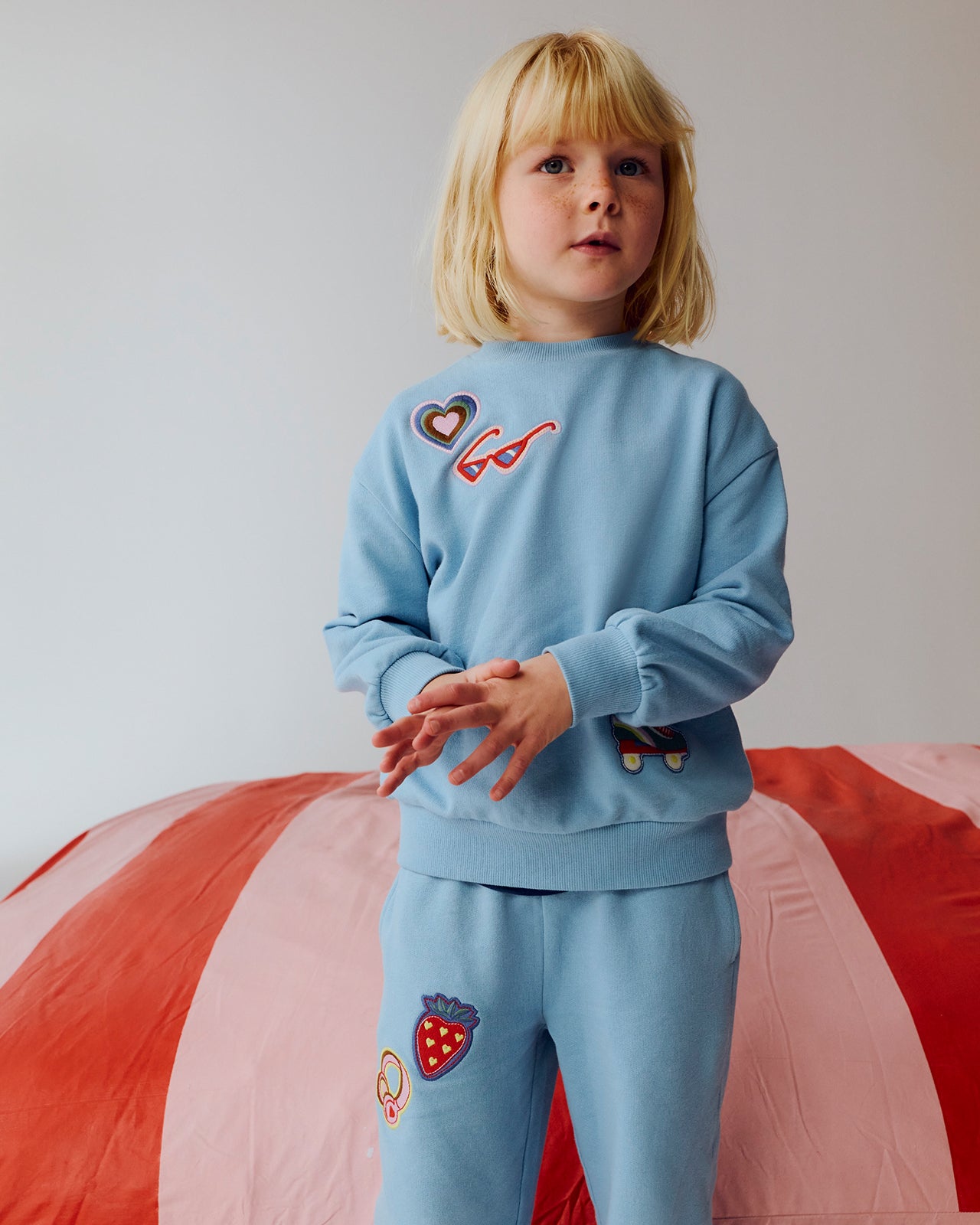 Child wearing a light blue sweatshirt and pants with colorful designs on a red and white striped background.