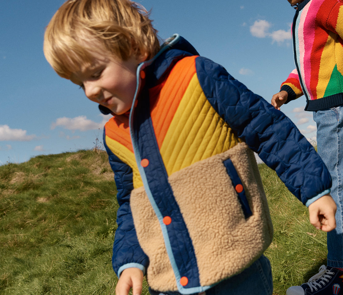Child wearing a colorful quilted jacket outdoors on a grassy hill with a blue sky.