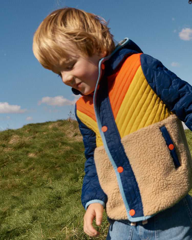 Child wearing a colorful quilted jacket outdoors on a grassy hill with a blue sky.