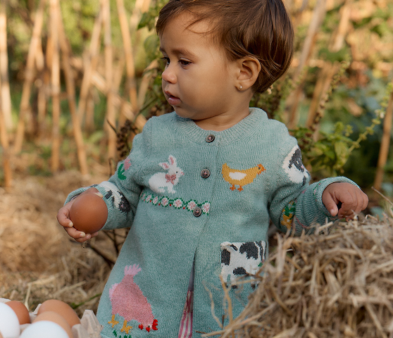Child holding a brown egg in a field with a knitted sweater featuring farm animal designs.