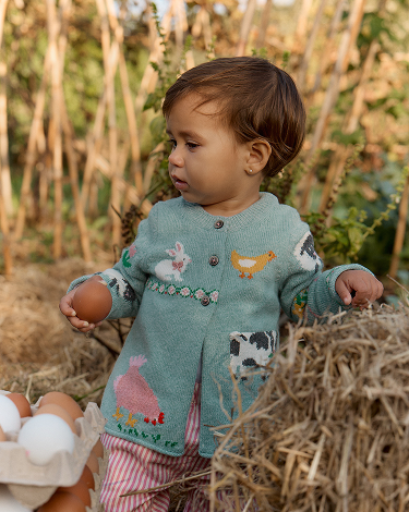 Child holding a brown egg in a field with a knitted sweater featuring farm animal designs.