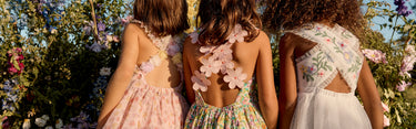 Three girls in floral dresses standing back-to-back in a garden.