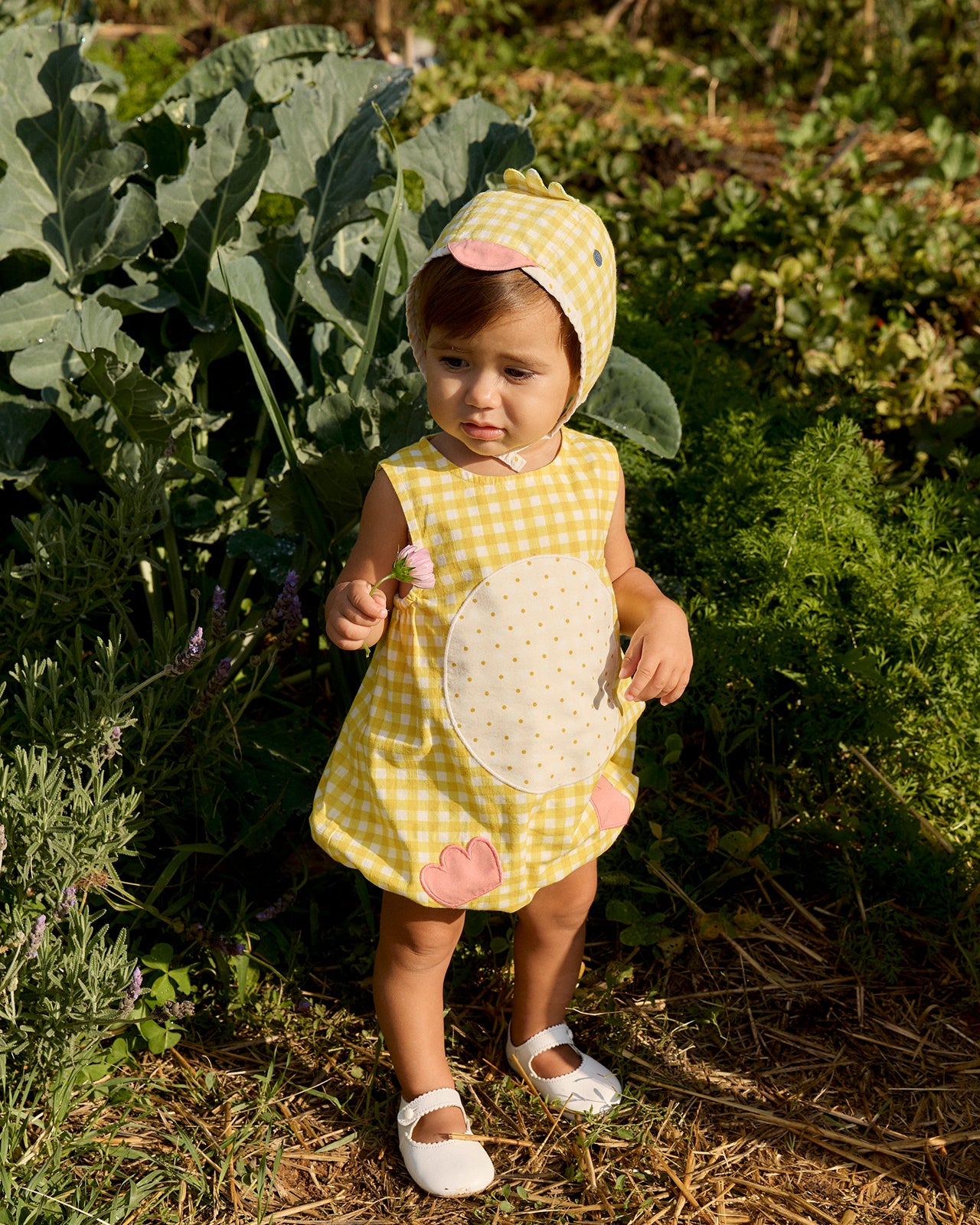 Child in a yellow checkered dress and bonnet standing in a garden