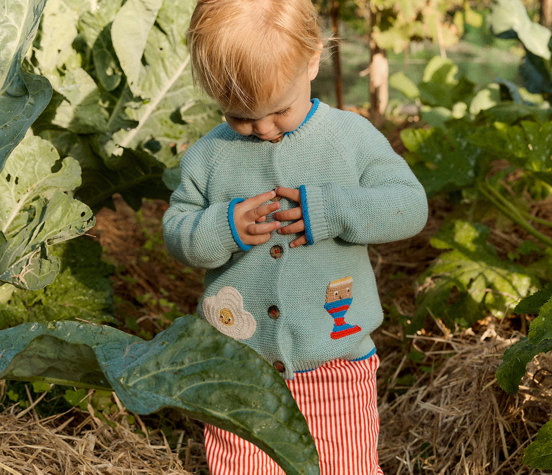 Child in a green cardigan and red striped pants standing among leafy plants.