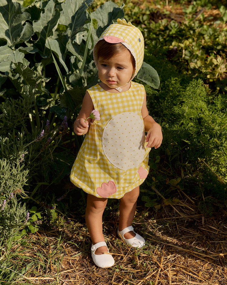 Child in a yellow checkered dress with a floral pattern standing in a garden.