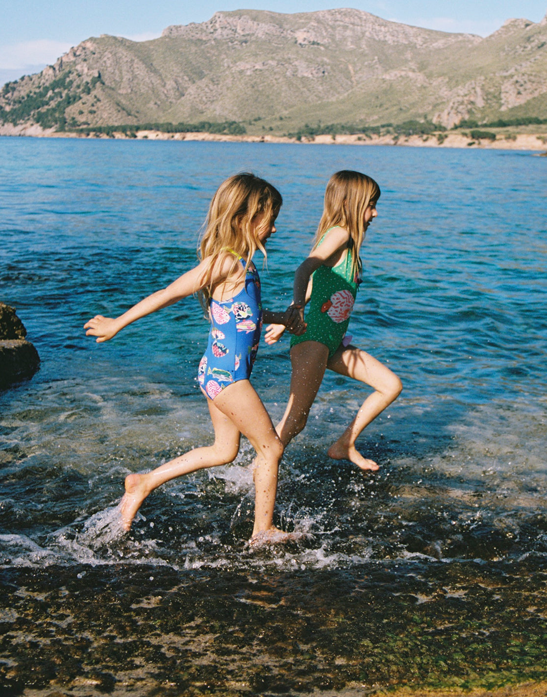 Girls running through the sea, holding hands and wearing fun Boden swimsuits
