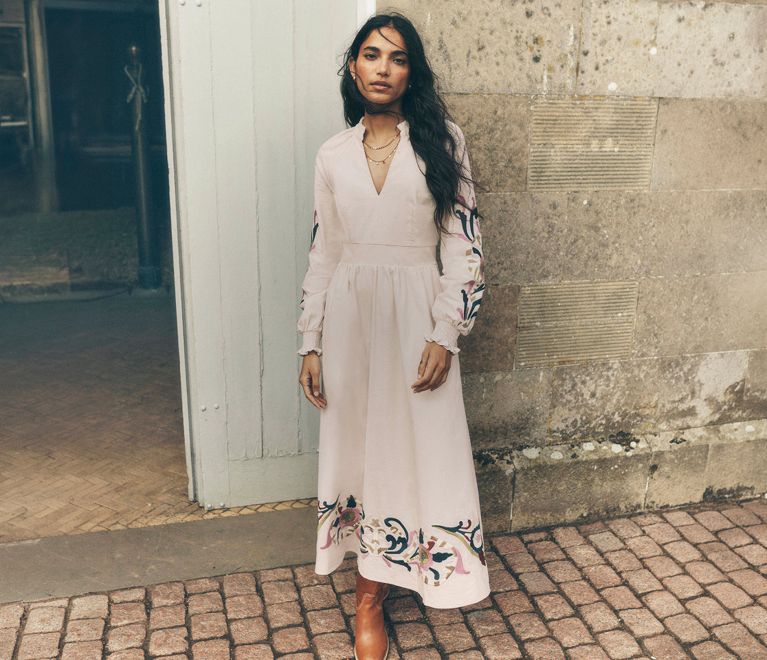 A woman stands outdoors wearing a long-sleeve midi dress in a very pale blush pink or off-white tone, with floral embroidery near the hem and sleeves. The background shows a stone wall, a doorway, and cobblestone pavement.