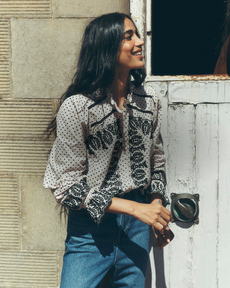 Person standing beside a weathered doorway, wearing a cream polka-dot blouse with intricate black embroidery and gathered sleeves, paired with high-waisted blue jeans. The image is set against a stone wall and distressed white wooden door, highlighting the detailed embroidery and relaxed silhouette.