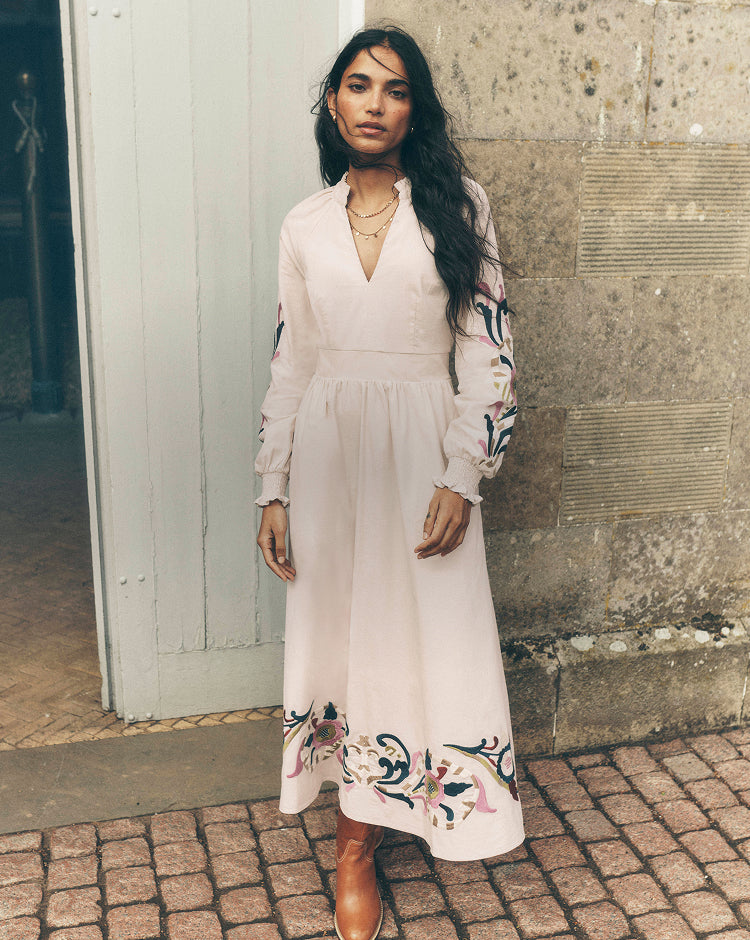 Person standing in a doorway wearing a light cream midi dress with colourful embroidered detailing on the sleeves and hem, paired with tan leather boots and layered gold necklaces. The look is set against a stone wall and cobbled pavement, creating a relaxed, heritage-inspired setting.