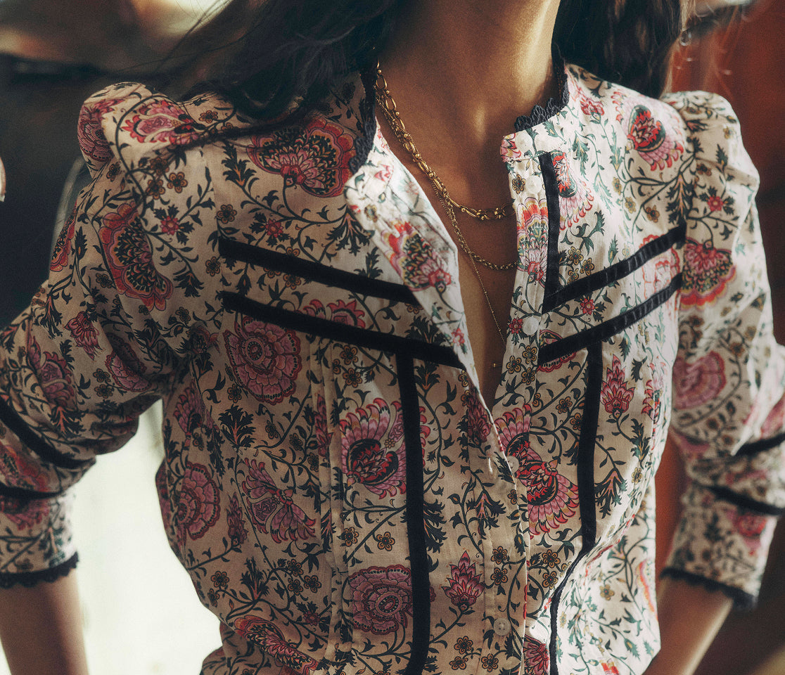 A close-up of a woman wearing a cream-colored blouse with a red and black floral paisley pattern, featuring black lace trim along the collar and front placket.