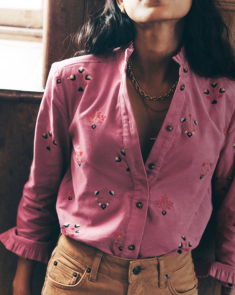 Close-up of a person wearing a pink button-front shirt with delicate multicoloured floral embroidery, paired with tan jeans and layered gold necklaces. The image is softly lit and focuses on the intricate embroidery, ruffled cuffs and textured fabric details.
