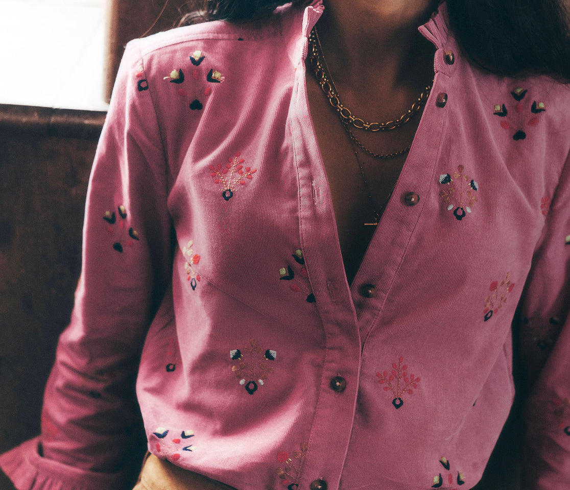Close-up of a person wearing a pink button-front shirt with delicate multicoloured floral embroidery, paired with tan jeans and layered gold necklaces. The image is softly lit and focuses on the intricate embroidery, ruffled cuffs and textured fabric details.