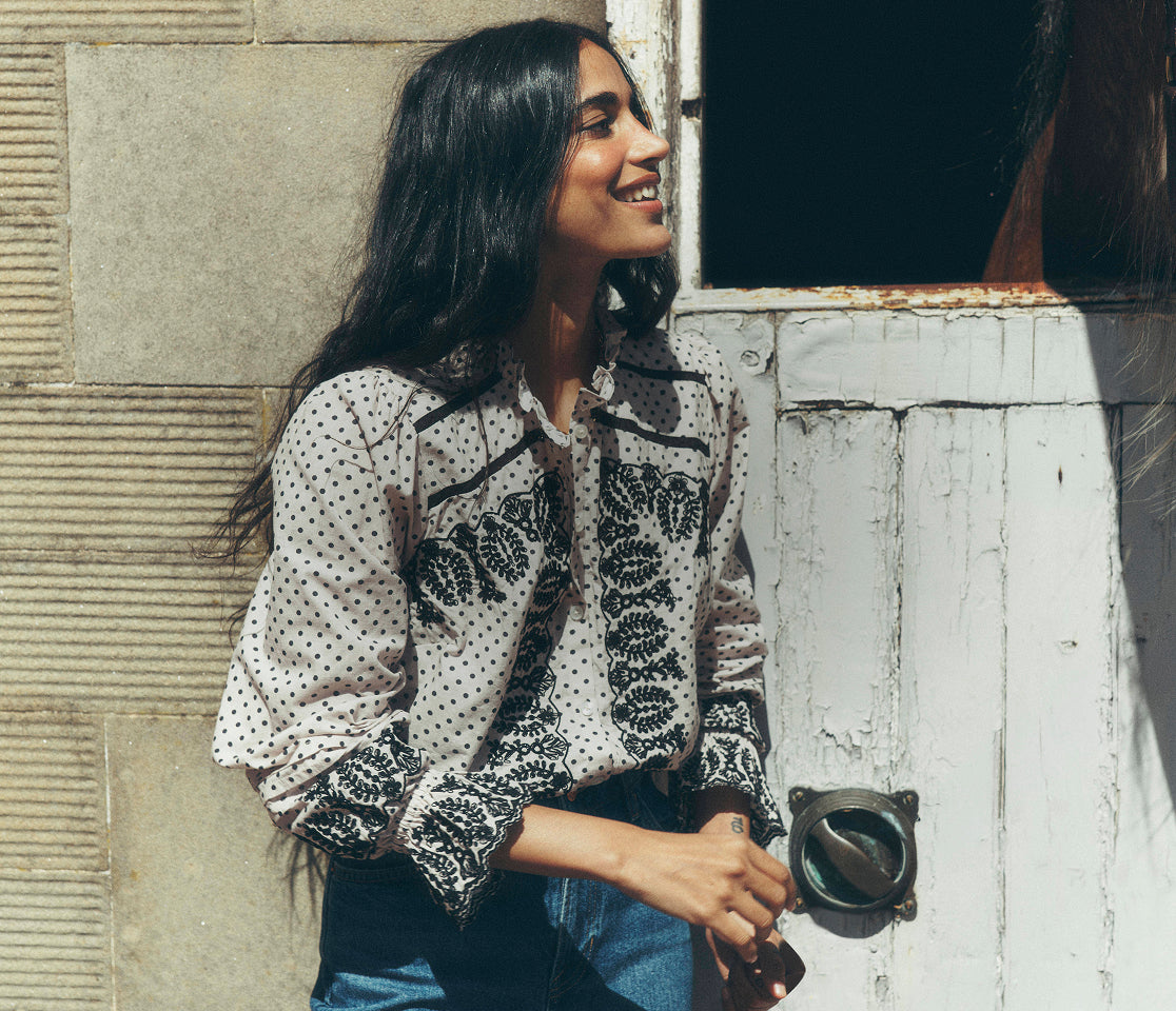 Person standing beside a weathered doorway, wearing a cream polka-dot blouse with intricate black embroidery and gathered sleeves, paired with high-waisted blue jeans. The image is set against a stone wall and distressed white wooden door, highlighting the detailed embroidery and relaxed silhouette.
