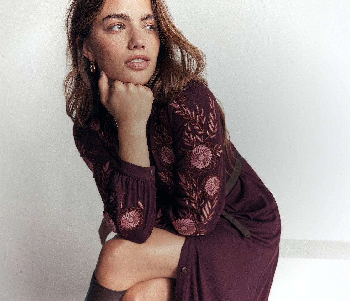 A woman wearing a dark burgundy long-sleeve dress with floral embroidery and sheer lace sleeves, posing in a studio setting against a light gray background.
