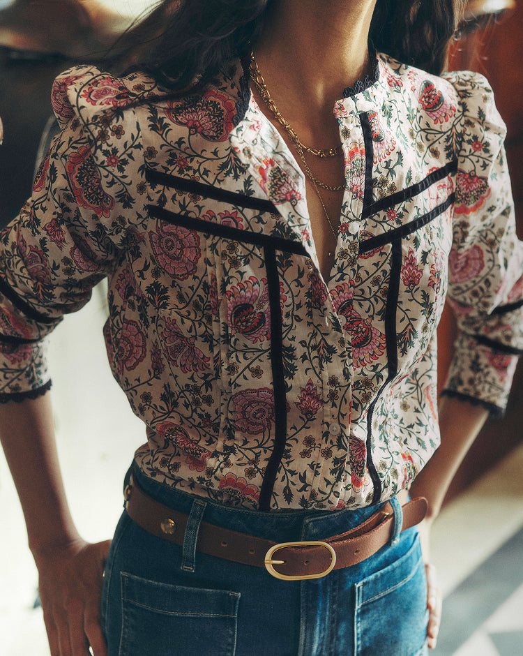 A close-up of a woman wearing a cream-colored blouse with a red and black floral paisley pattern, featuring black lace trim along the collar and front placket.