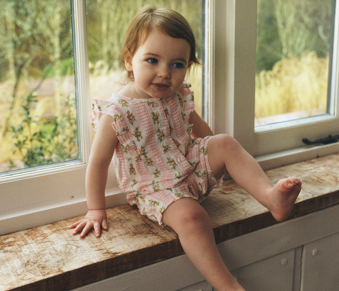 Child in a floral outfit sitting on a windowsill with a garden view.