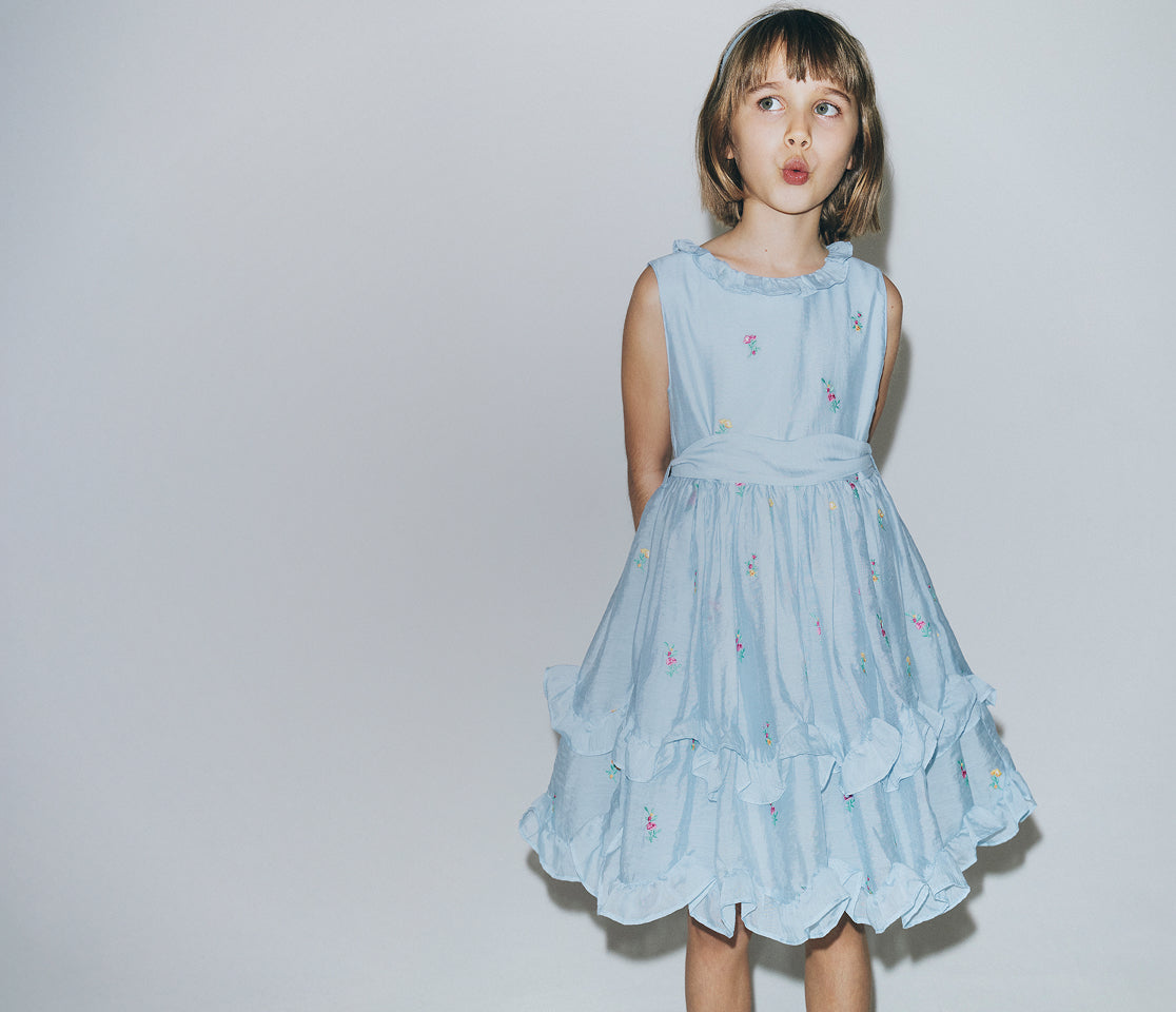 Young girl wearing a light blue dress with floral details on a plain background