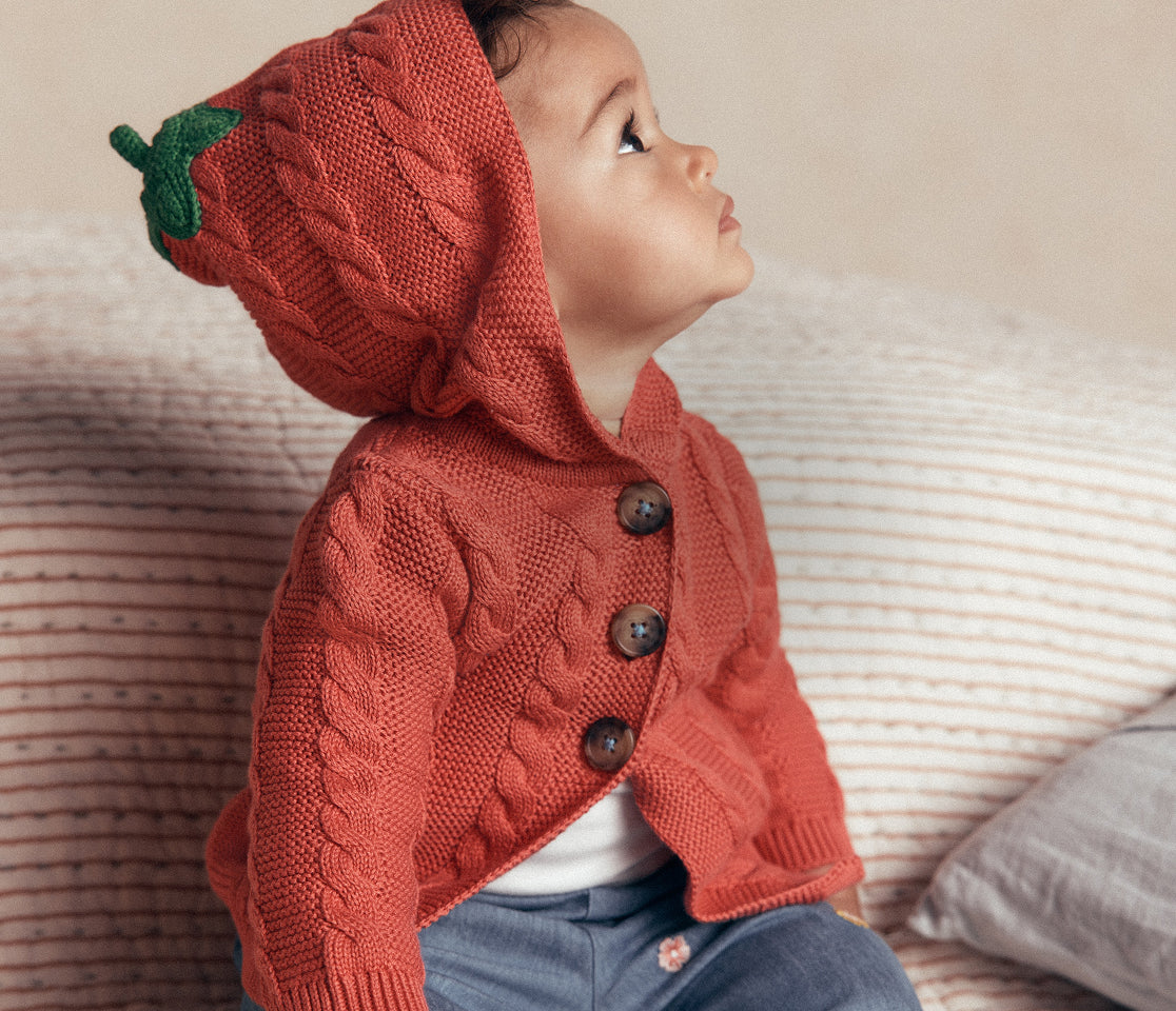 A baby wearing a knitted hooded cardigan styled like a strawberry, featuring a textured cable-knit body, dark buttons, and a green leaf detail on the hood, posed on a striped couch.