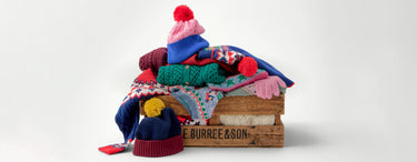 Stack of colorful winter hats and scarves on a wooden box with 'Burree & Son' branding.
