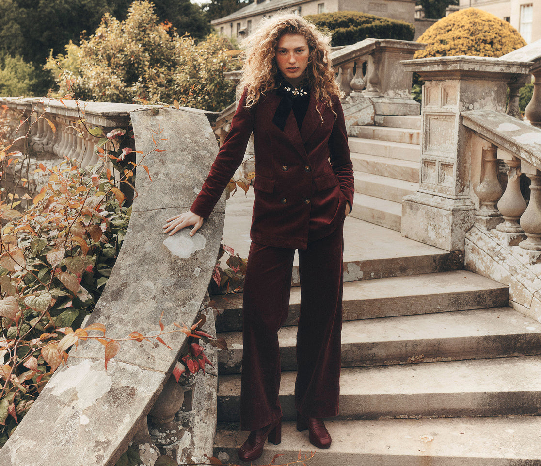 standing on stone steps in a formal garden setting, wearing a deep burgundy double-breasted velvet blazer with matching wide-leg trousers and platform shoes. A jeweled collar detail adds embellishment to the tailored look, while the historic stone balustrades and autumn foliage create an elegant heritage-inspired backdrop.