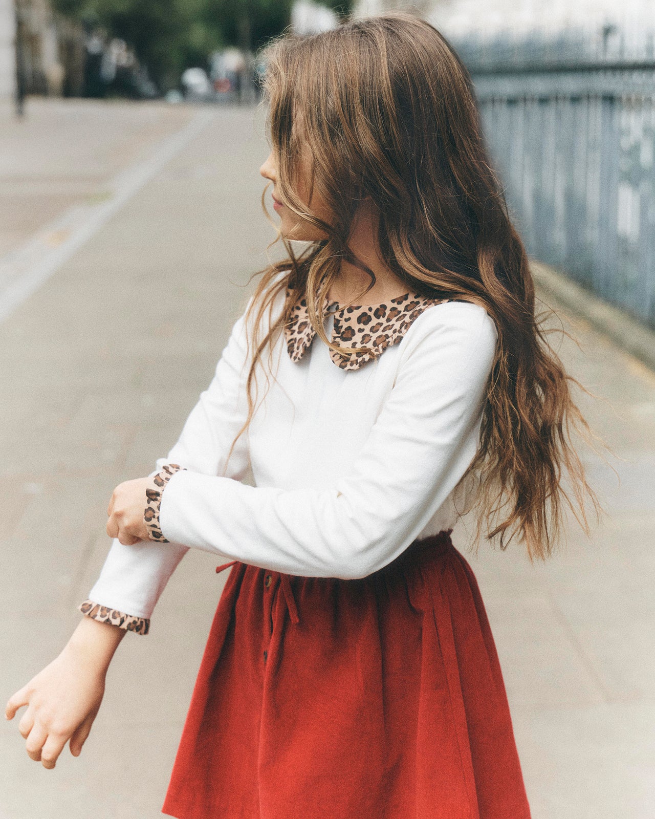 A young girl with long brown hair is shown from a side-back angle outdoors, wearing a white long-sleeve top with a leopard-print collar and cuffs, paired with a rust-red skirt.