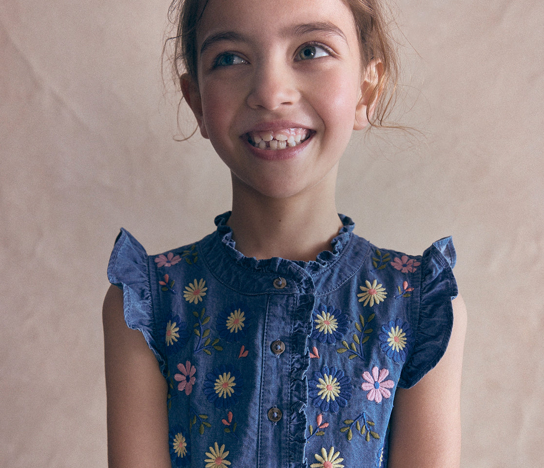 A young girl smiling and looking to the side, wearing a blue denim-style top with colorful embroidered flowers and ruffled sleeves, against a plain beige background.