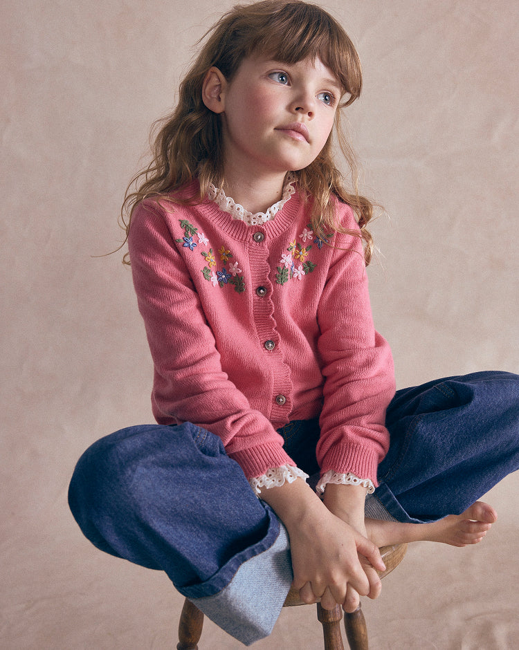 A young girl with light brown hair sits cross-legged on a wooden stool against a textured beige studio backdrop. She is wearing a pink button-front cardigan with colorful floral embroidery and lace trim, paired with dark blue pants.