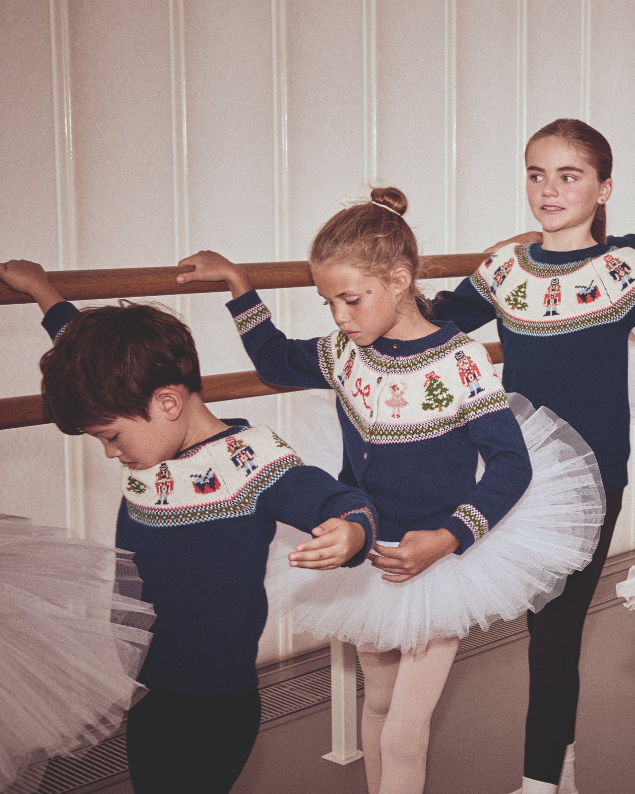 Three children in ballet attire with festive sweaters practicing ballet in a studio.