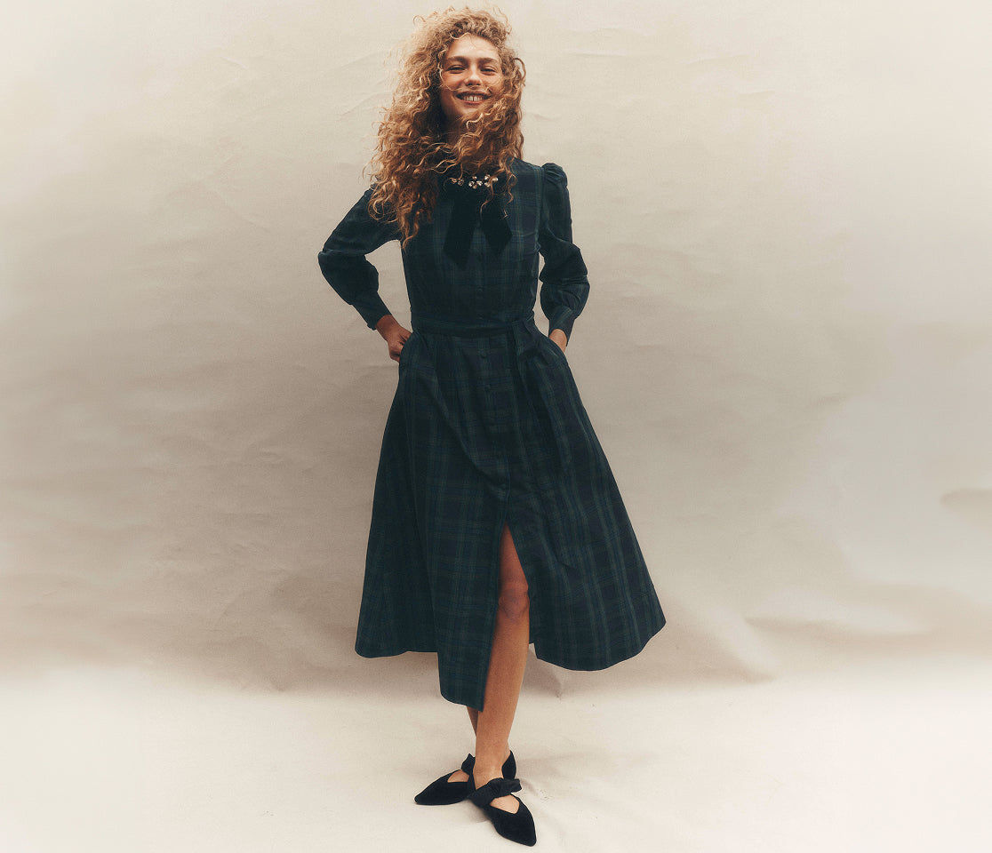 Women standing in a studio setting wearing a dark green and navy tartan midi dress with long sleeves, a button-front design and a fitted waist. The dress is styled with a jeweled collar embellishment and black bow-detail flats, with the full silhouette shown against a soft neutral backdrop.