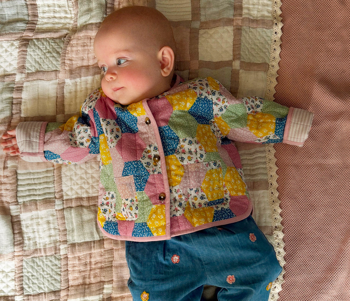 A baby lying on a quilted blanket wearing a colorful patchwork quilted cardigan and blue pants.